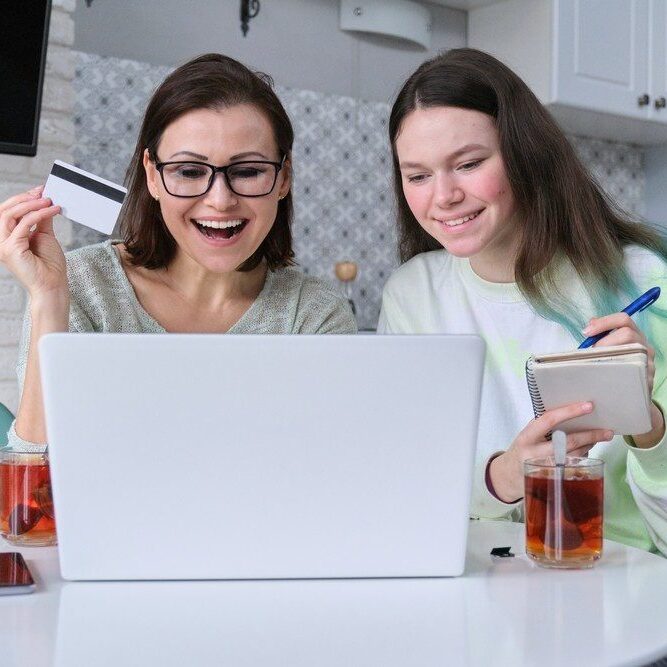 Family, mother and teenage daughter using laptop computer credit card and doing online shopping. Women sitting at home in kitchen talking smiling choosing gifts